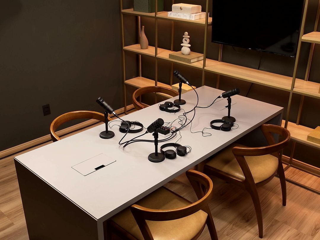 Mesa de podcast do Estúdio B · M8 vista em diagonal, com microfones, fones de ouvido e cadeiras de madeira diante da estante iluminada
