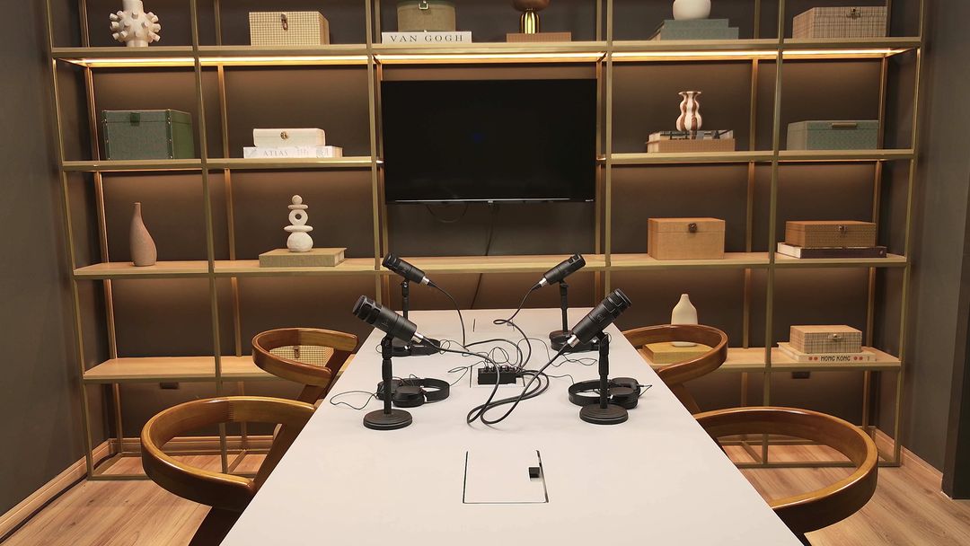 Set de podcast do Estúdio B · M8, com mesa branca, quatro microfones com fones de ouvido e a estante iluminada ao fundo